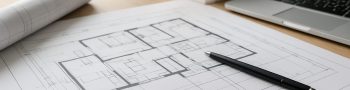 Blueprint of a residential house plan on a wooden table with a model house and a laptop in the background