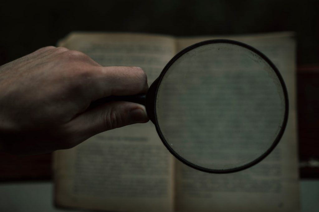 A hand holding a magnifying glass over an open book with blurred text in the background.