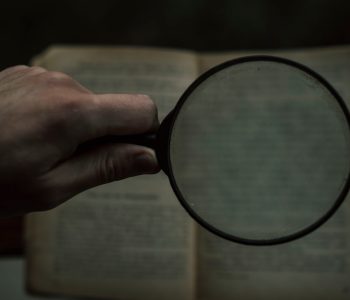 A hand holding a magnifying glass over an open book with blurred text in the background.