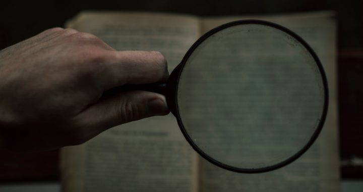 A hand holding a magnifying glass over an open book with blurred text in the background.
